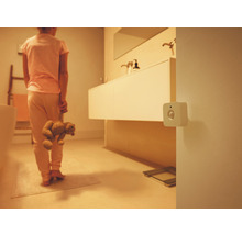 En vit kvadratisk rörelsedetektor på väggen i ett dämpat belyst badrum med en person som håller i en nallebjörn.