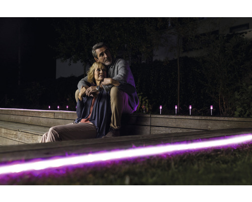 Ett par sitter på en träterrass på kvällen, som är upplyst med violetta lampor. Gräsmatta och buskar syns i bakgrunden.