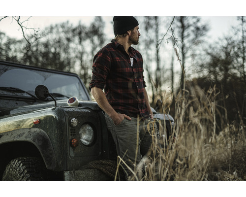 En man i rutig skjorta lutar sig mot ett terrängfordon i en lantlig miljö.