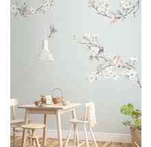 Matsal med ljust träbord, stolar, pall, porslin, krukväxt och en väggtapet med fågel- och blommotiv.