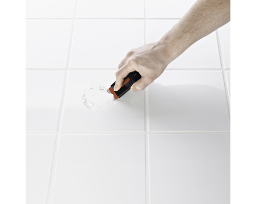 Hand med fogskrapa tar bort fogmassa mellan plattor.