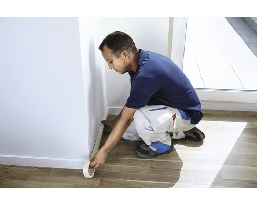 Man tejpar maskeringstejp på en vägg längs en golvlist.