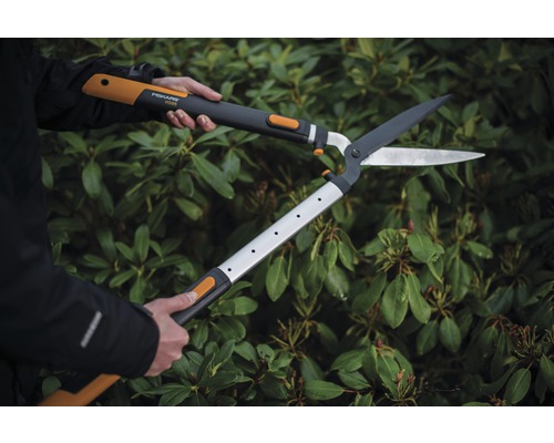 En person använder en häcksax med utväxling framför en grön häck.