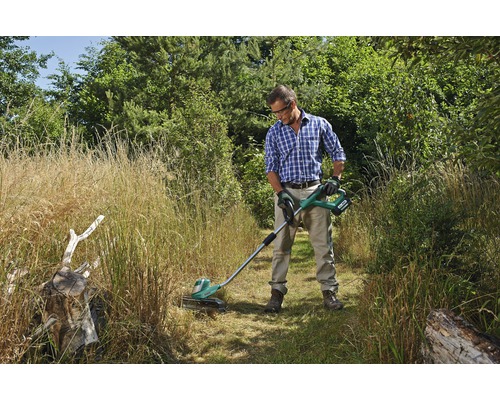 En man använder en batteridriven grästrimmer i en trädgård med högt gräs.