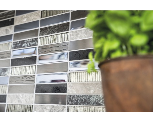 Detaljbild av en vägg med mosaikplattor i olika gråtoner, bredvid en växt