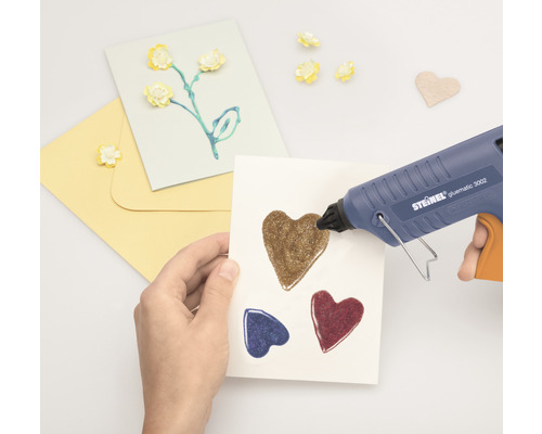 Hand som limmar hjärtan på ett kort med en limpistol. Hantverksmaterial ligger bredvid.