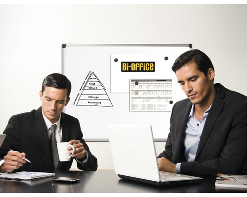 Två män sitter vid ett bord med en bärbar dator framför en whiteboardpresentation på ett kontor.