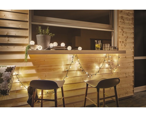 Träterrass med bar, upplyst med ljusslingor och dekorerad med växter och drycker, kompletterad med barstolar.