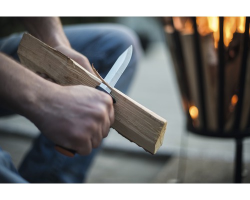 En person täljer trä med en kniv framför en eldfat.