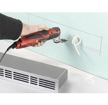 En person använder ett multifunktionsverktyg för att skära ett rör på en vägg nära ett element