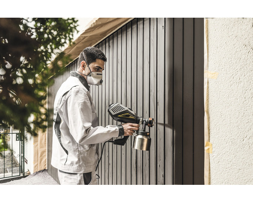 Man med andningsskydd sprutar färg på trästaket med färgspruta