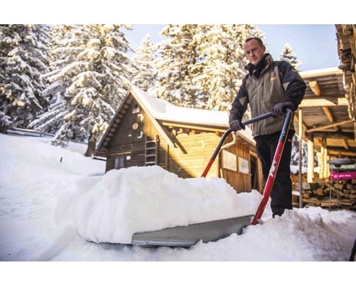 Man skottar snö med en snöskyffel framför ett trähus.