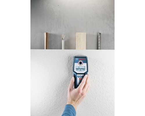 En hand håller en Bosch detektor mot en vägg. På en hylla i bakgrunden står en kopparledning, en elkabel, en träbit och en armering.