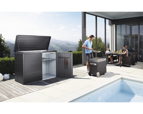 Trädgårdsscen med redskapsbod, grill och familj vid matbordet på terrassen