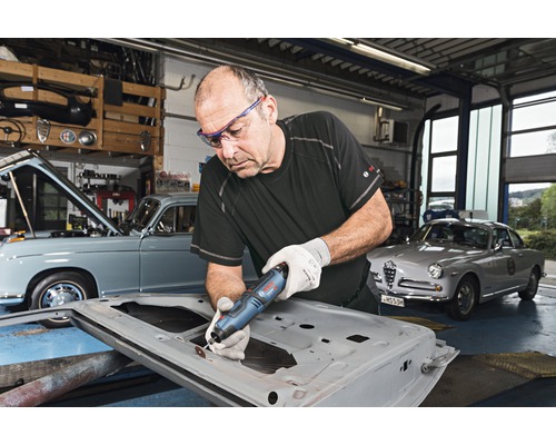 Man arbetar med ett multifunktionsverktyg på en bildörr i en verkstad med veteranbilar.