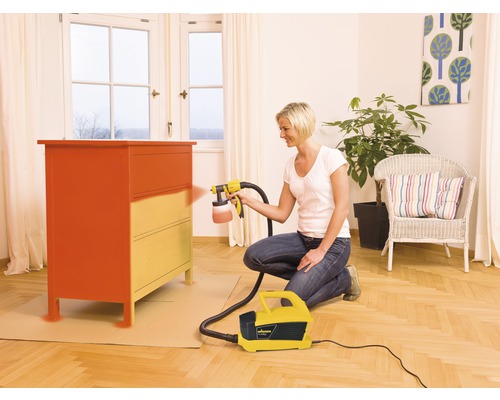 Kvinna lackerar byrå med färgspraysystem i ett vardagsrum.