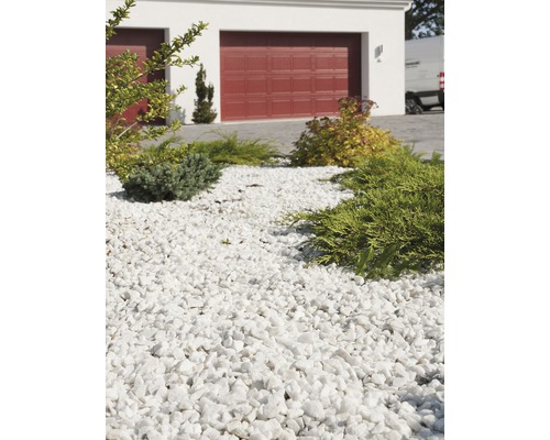 Dekorativ trädgård med vita marmorkross och växter framför ett hus med garageport.