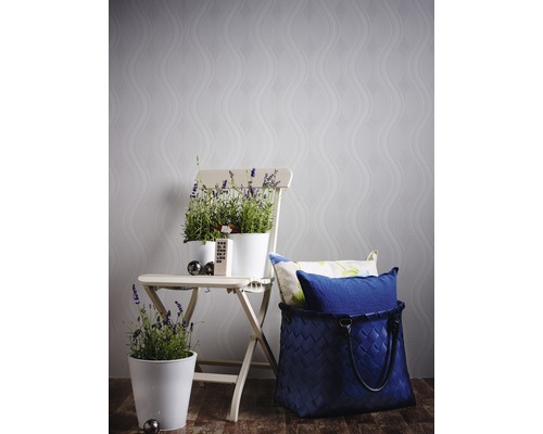 Dekorativ scen med blomkrukor på en hopfällbar stol framför en mönstrad tapet och en flätad korg med kuddar.