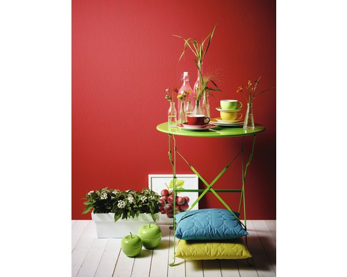 Dekorativ scen med ett grönt trädgårdsbord, porslin, växter, dekoration och kuddar mot en röd vägg.