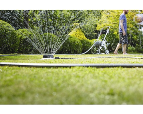 Grässpridare vattnar en gräsmatta med en slangvagn och en person i bakgrunden.