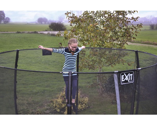 Flicka balanserar på en trädgårdstrampolin med skyddsnät. EXIT Logo.