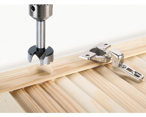 Forstnerborr borrar hål bredvid ett gångjärn i en träskiva.