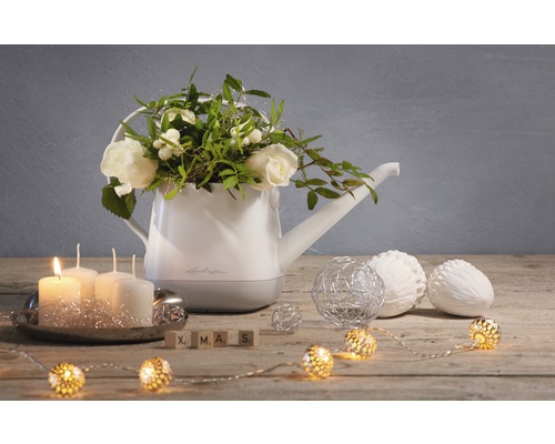 Dekorativ vattenkanna med blomsterarrangemang, ljus, kulor och ljusslinga på ett träbord