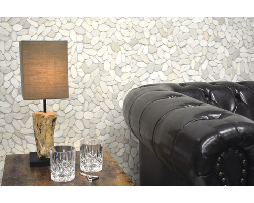 Miljöbild med en bordslampa på ett träbord, två glas, en kapsylöppnare och en lädersoffa framför en vägg med stenar.