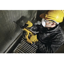 En arbetare borrar ett hål i en betongvägg med en borrhammare. Han bär skyddskläder som skyddsglasögon, hjälm och handskar.