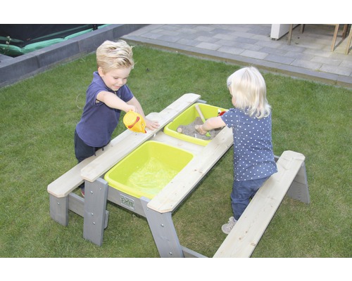 Två barn leker vid ett grått picknickbord med sand och vatten i gröna behållare.