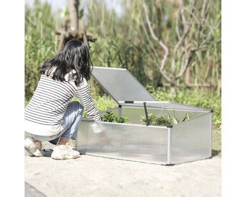 Kvinna böjer sig ner mot en odlingskrage med öppet lock i trädgården.