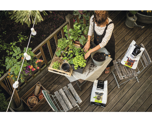 Kvinna planterar växter på en träterrass med planteringsjord, omgiven av trädgårdsredskap och dekorationer