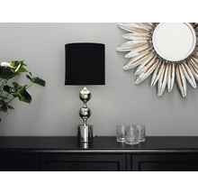 Dekorativ scen med en bordslampa med svart skärm och förkromad fot, en spegel i fjäderdesign och glas på ett svart skåp.