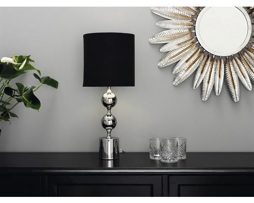 Dekorativ scen med en bordslampa med svart skärm och förkromad fot, en spegel i fjäderdesign och glas på ett svart skåp.