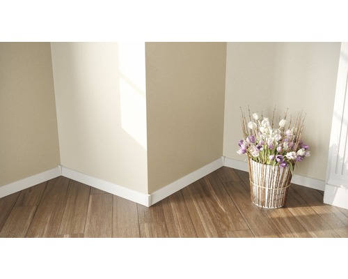 Hörn av ett ljust rum med trägolv, vita golvlister och en bukett i en kruka