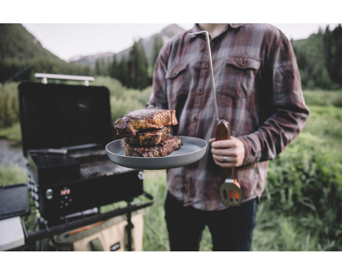 Man med grill, stekspade och tallrik med biffar utomhus