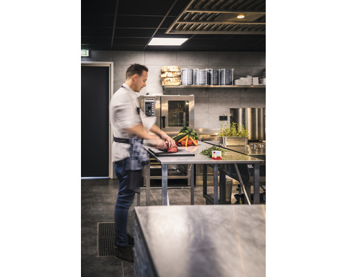 En kock förbereder grönsaker och kött på ett rostfritt stålbord i ett professionellt kök.