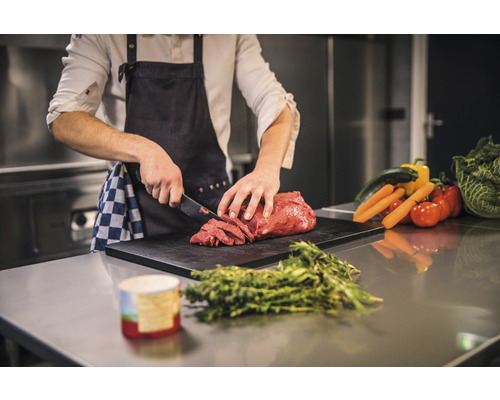En person skär kött på en skärbräda med grönsaker i bakgrunden.