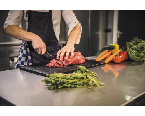 En person skär kött på en skärbräda, omgiven av grönsaker och örter.