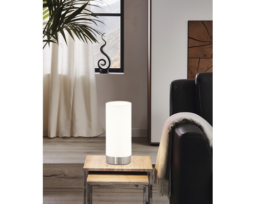 Miljöbild med en cylinderformad bordslampa i glas och metall på ett träbord