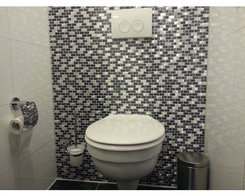 Badrum med toalett, toalettpappershållare, toalettborste och papperskorg framför en vägg med mosaikplattor