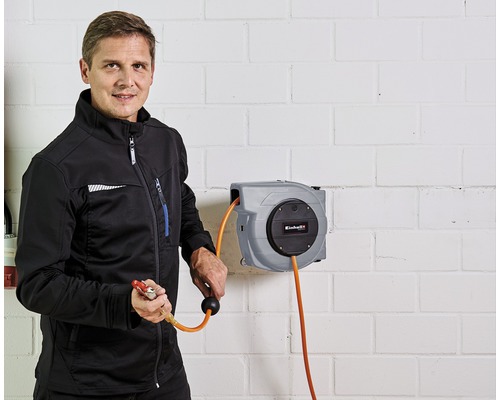 En man håller en luftpistol vid en Einhell luftslangrulle på en tegelvägg