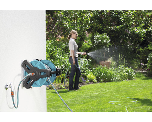 Trädgårdsscen med väggmonterad slangvinda och kvinna som vattnar en trädgård