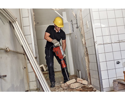 En man med skyddshjälm och skyddsglasögon arbetar med en bilningshammare i ett kaklat rum.