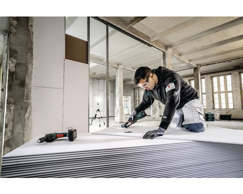 En hantverkare skär en gipsskiva med ett specialverktyg i ett oinrett hus, med andra verktyg redo.