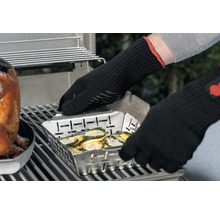 På en grill står en metallgrillpanna med grönsaker, som hålls av någon med grillhandskar.