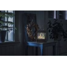 Dekorativ snöglobslykta med vinterlandskap på ett bord, omgiven av växter och en krans.