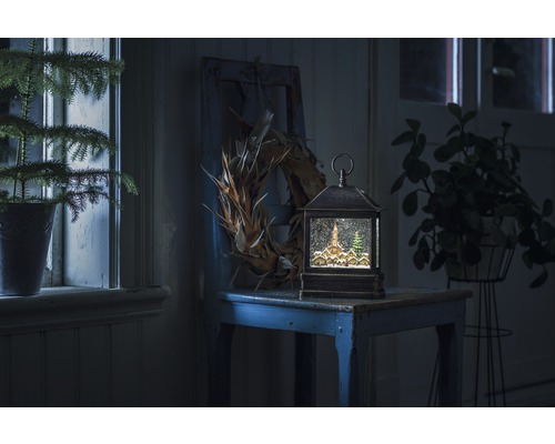 Dekorativ snöglobslykta med vinterlandskap på ett bord, omgiven av växter och en krans.