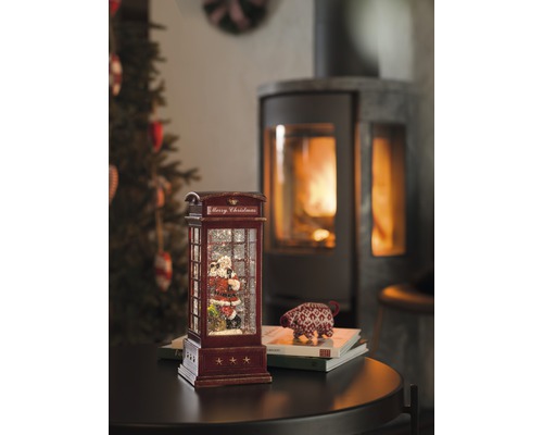 Dekorativ snöglob i form av en telefonkiosk med jultomtemotiv och öppen spis i bakgrunden.