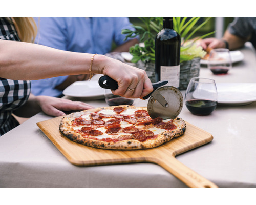 Scen med en person som skär en pizza med en pizzaslicer på en träbräda, med en flaska vin och glas i bakgrunden, på ett bord utomhus